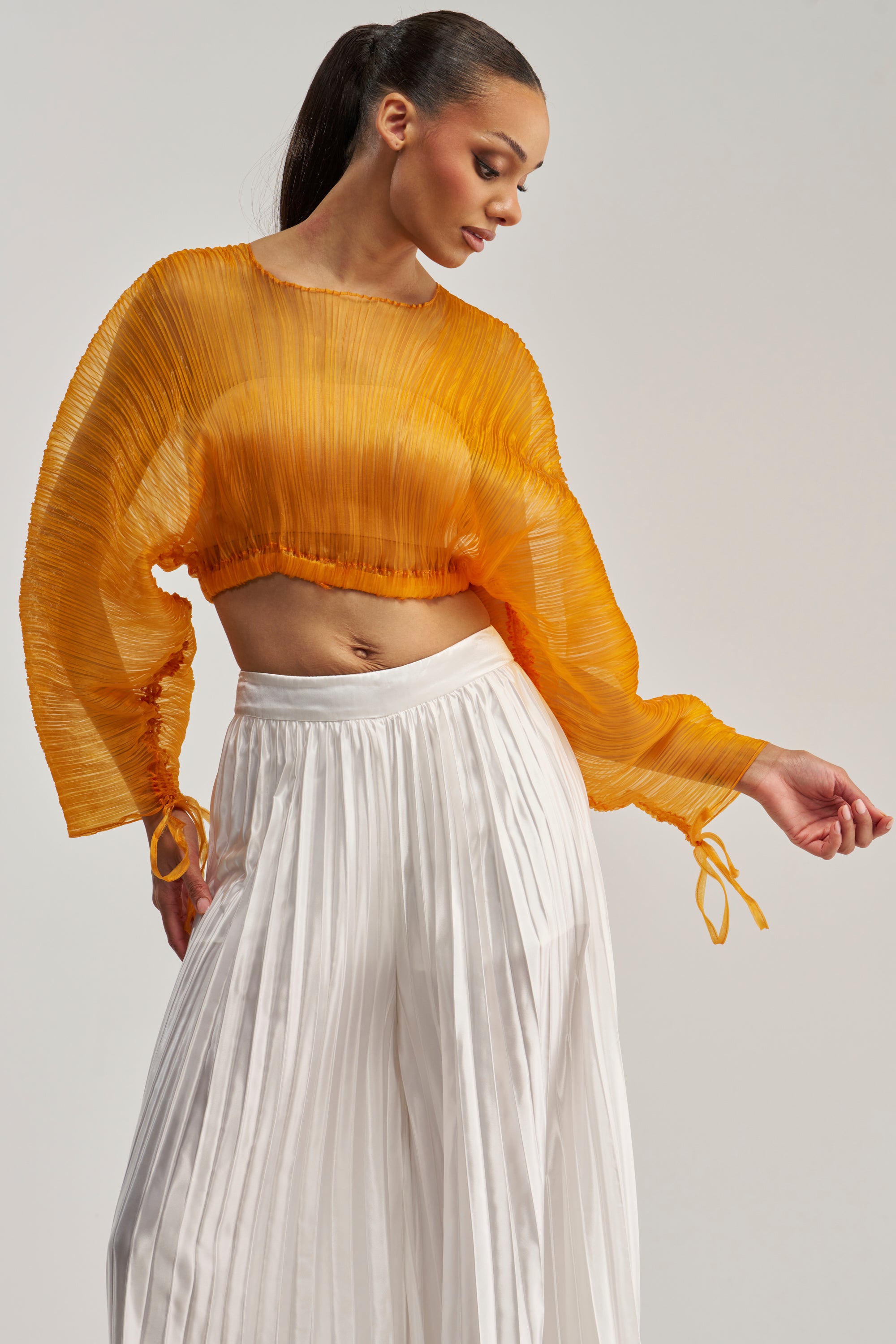 A woman poses against a neutral background, wearing the FORTUNES FAVORITE SHEER LONG SLEEVE BLOUSE in marigold paired with high-waisted, white wide-leg pleated trousers.