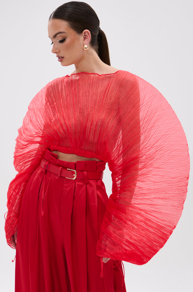 A woman in the bold, bright red FORTUNE'S FAVORITE ORGANZA CROP TOP with wide, pleated fan sleeves and high-waisted belted pants looks down, her hair in a sleek ponytail against a plain background—she’s Fortune’s Favorite.