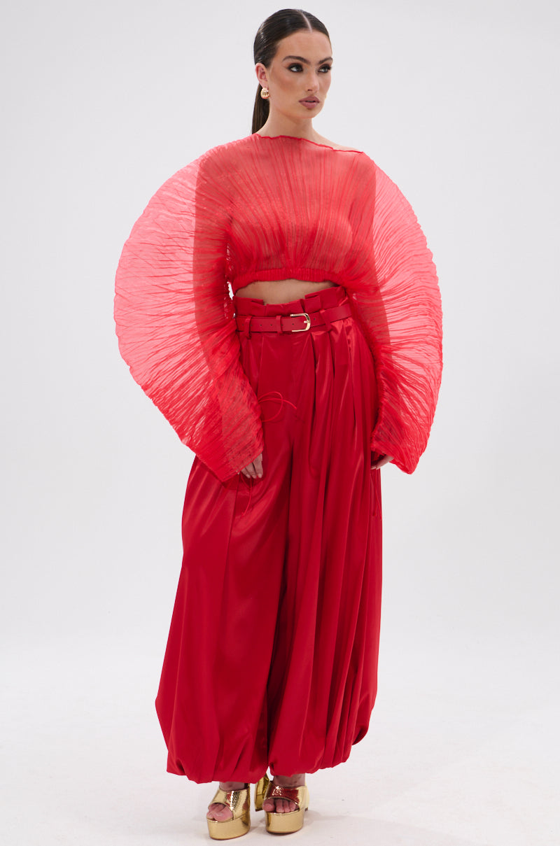A woman stands against a white background wearing the FORTUNE'S FAVORITE ORGANZA CROP TOP IN RED with wide high-waisted pants, sleek hair, hoop earrings, and gold platform shoes.