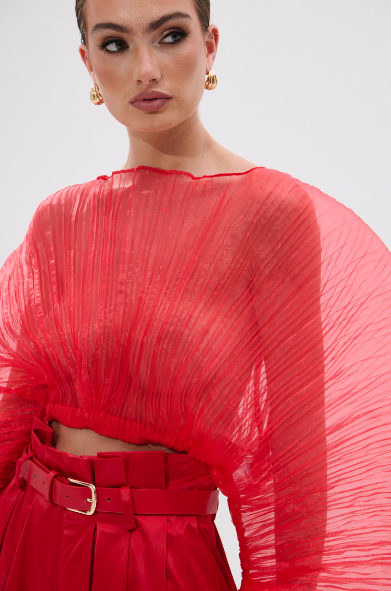 A woman in the FORTUNE'S FAVORITE ORGANZA CROP TOP IN RED—sheer, pleated, and voluminous—pairs it with high-waisted belted pants, styling her slicked-back hair with gold hoop earrings.
