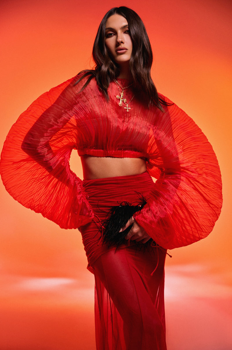 A woman with long, wavy dark hair poses against an orange background in the FORTUNE'S FAVORITE ORGANZA CROP TOP IN RED with sculptural pleated sleeves, paired with a matching red skirt and holding a black feathered accessory.