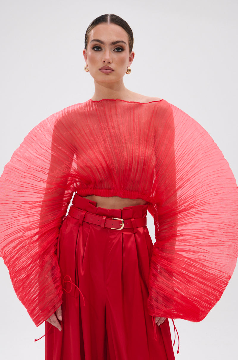 A woman wears the FORTUNE'S FAVORITE ORGANZA CROP TOP IN RED, featuring sheer pleats and large fan-like sleeves, paired with high-waisted red trousers and a wide belt, standing against a plain light background.
