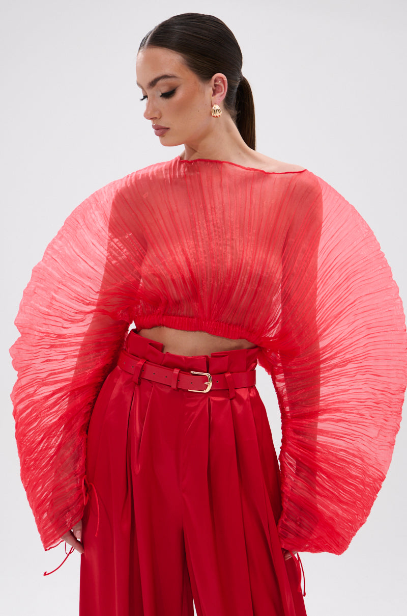 A woman in the FORTUNE'S FAVORITE ORGANZA CROP TOP IN RED, featuring dramatically wide sleeves and paired with high-waisted red belted pants, stands against a plain light background, looking down and to the side.