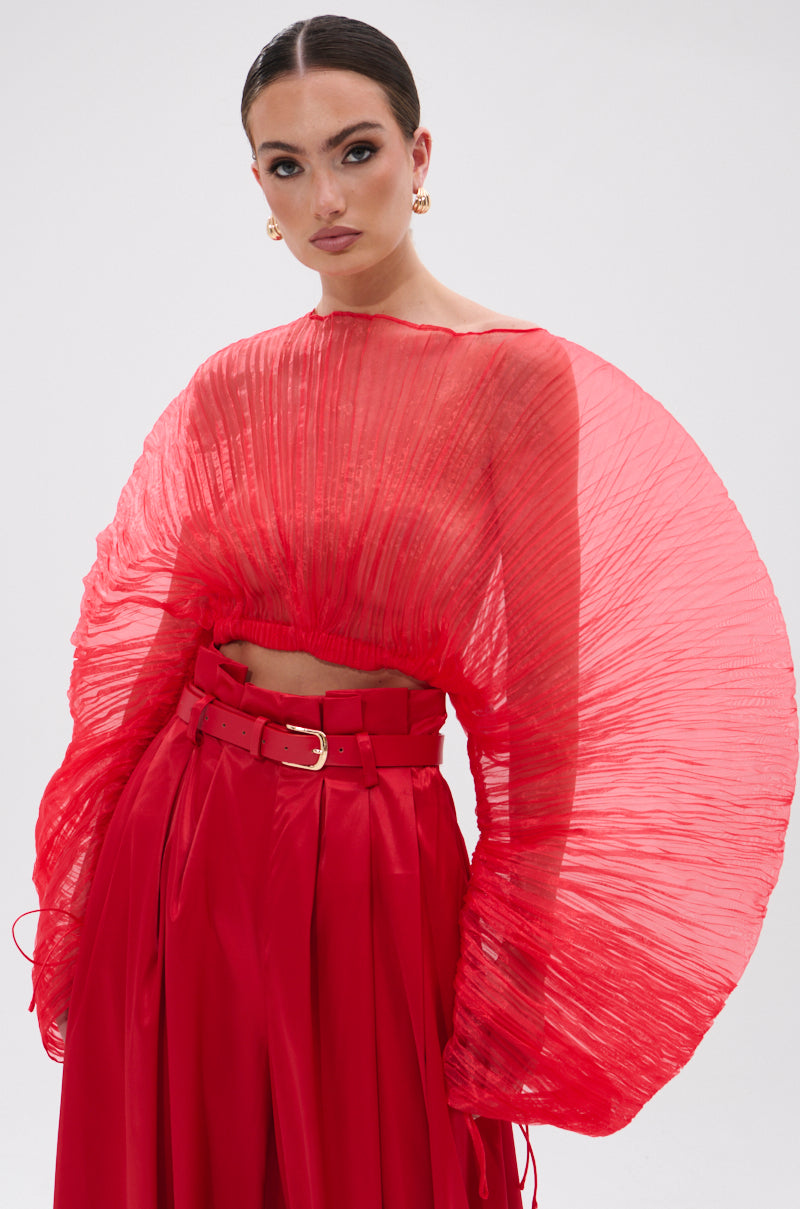 A woman poses in the FORTUNE'S FAVORITE ORGANZA CROP TOP IN RED, styled with high-waisted belted pants, dramatic pleated sheer sleeves, slicked-back hair, hoop earrings, and neutral makeup—truly Fortune’s Favorite.