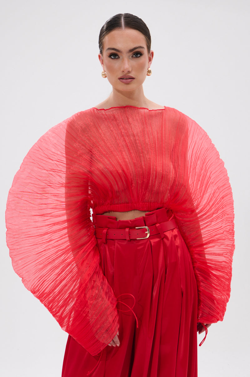 A woman wears the FORTUNE'S FAVORITE ORGANZA CROP TOP IN RED, a dramatic sheer red top with voluminous sleeves, paired with a matching skirt. With slicked-back hair, gold earrings, and bold makeup, she poses confidently against a white background.