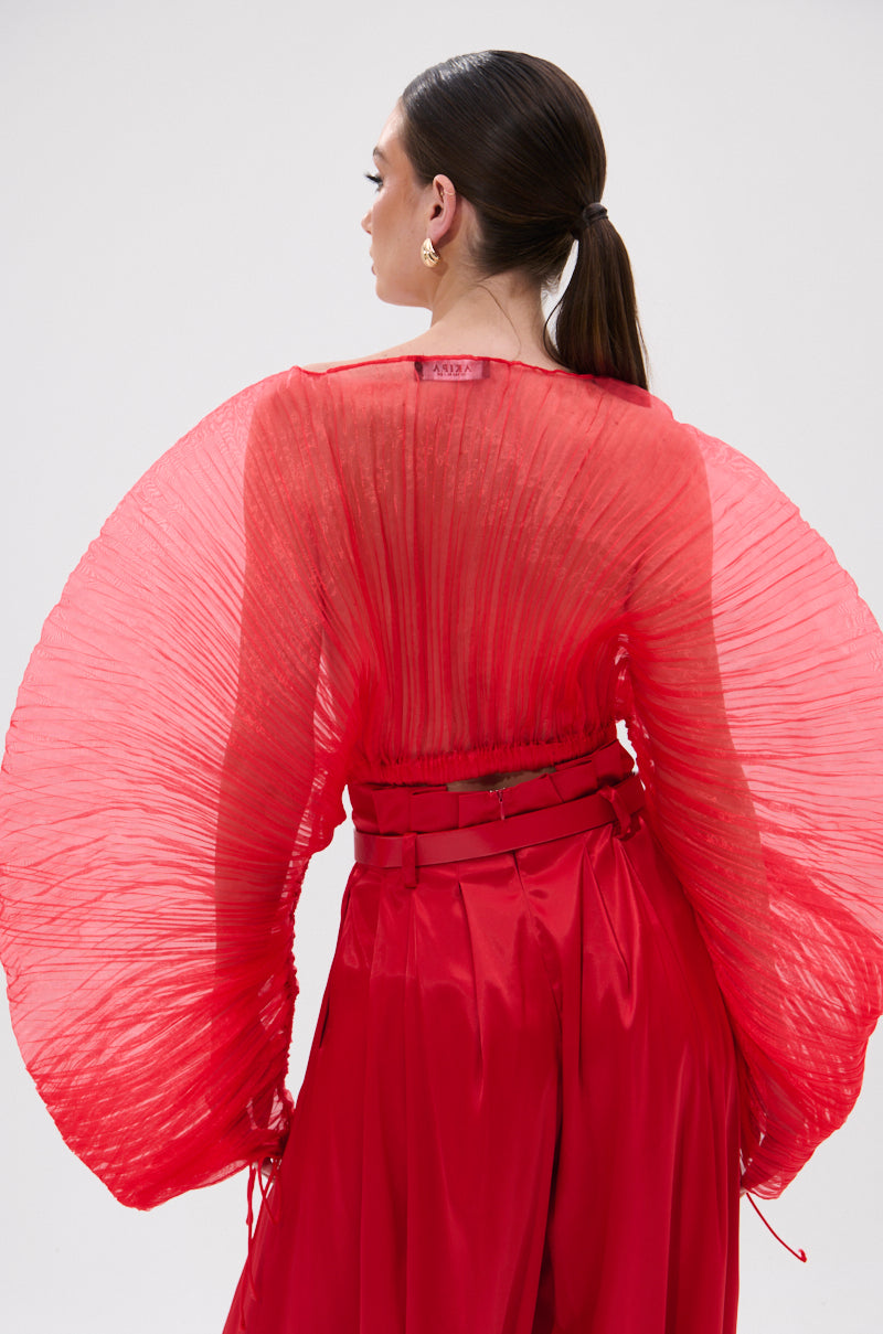 A woman with slicked-back hair models the FORTUNE'S FAVORITE ORGANZA CROP TOP IN RED, featuring sheer fabric, dramatic pleated wing sleeves, and a unique back detail as she faces away. She pairs it with matching high-waisted red pants.
