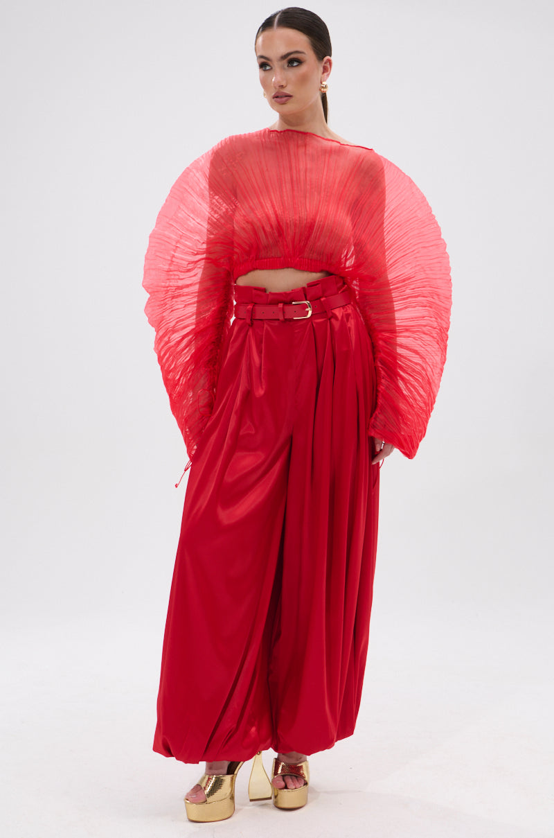 A woman poses against a plain background in the FORTUNE'S FAVORITE ORGANZA CROP TOP IN RED—sheer, pleated, and fan-shaped—styled with wide-legged red pants and gold platforms. Sleek hair and bold makeup complete her striking look.