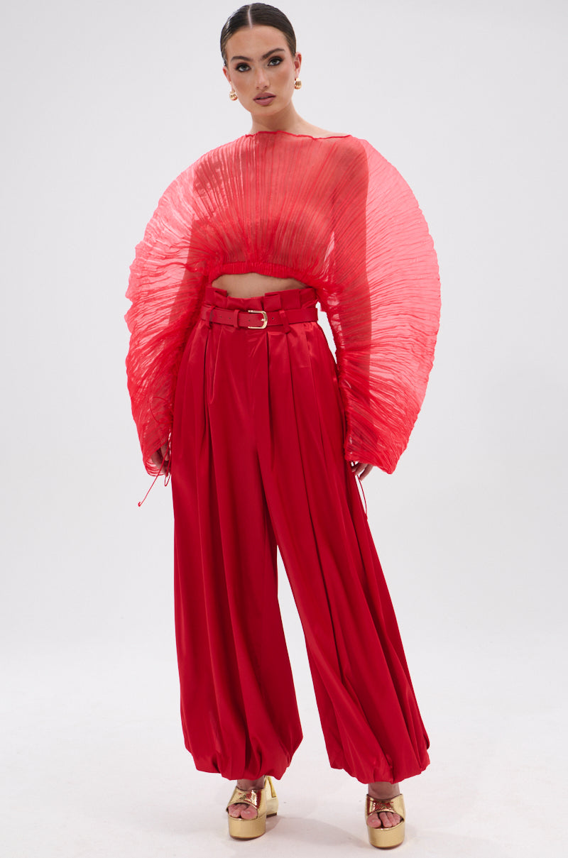 A woman in the FORTUNE'S FAVORITE ORGANZA CROP TOP IN RED—featuring wide pleated sleeves—pairs it with high-waisted loose red pants and gold platform shoes, standing against a plain white background.