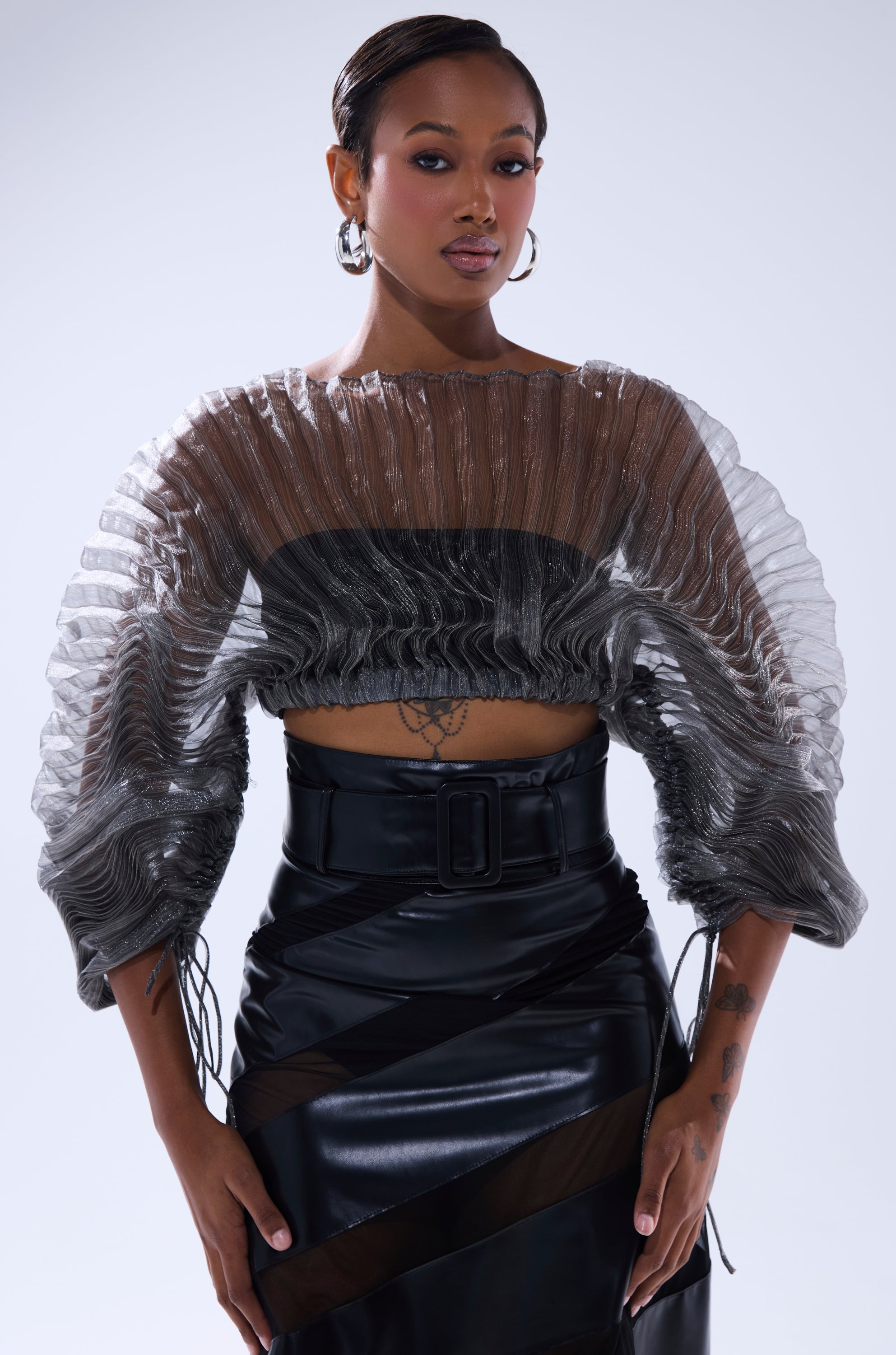 A woman poses confidently in the SHEER PLEATED CROP TOP IN GREY, styled with a high-waisted black faux leather skirt, hoop earrings, and slicked-back hair. Voluminous sleeves add a trendy touch to her look. 0