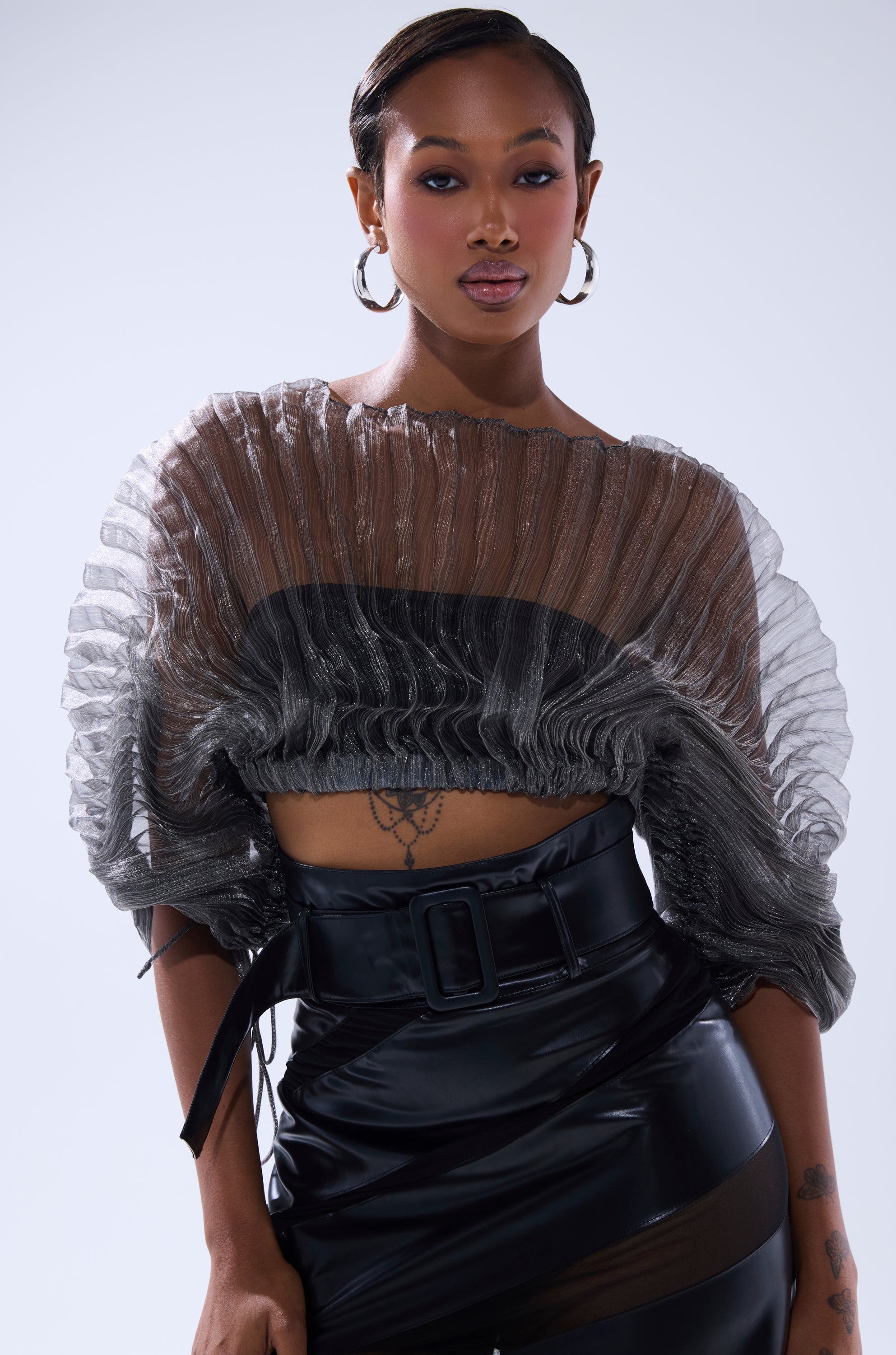A woman with short hair wears hoop earrings, a SHEER PLEATED CROP TOP IN GREY, and high-waisted black vinyl pants with a big belt. She stands confidently against a light background, revealing a tattoo on her torso. 1