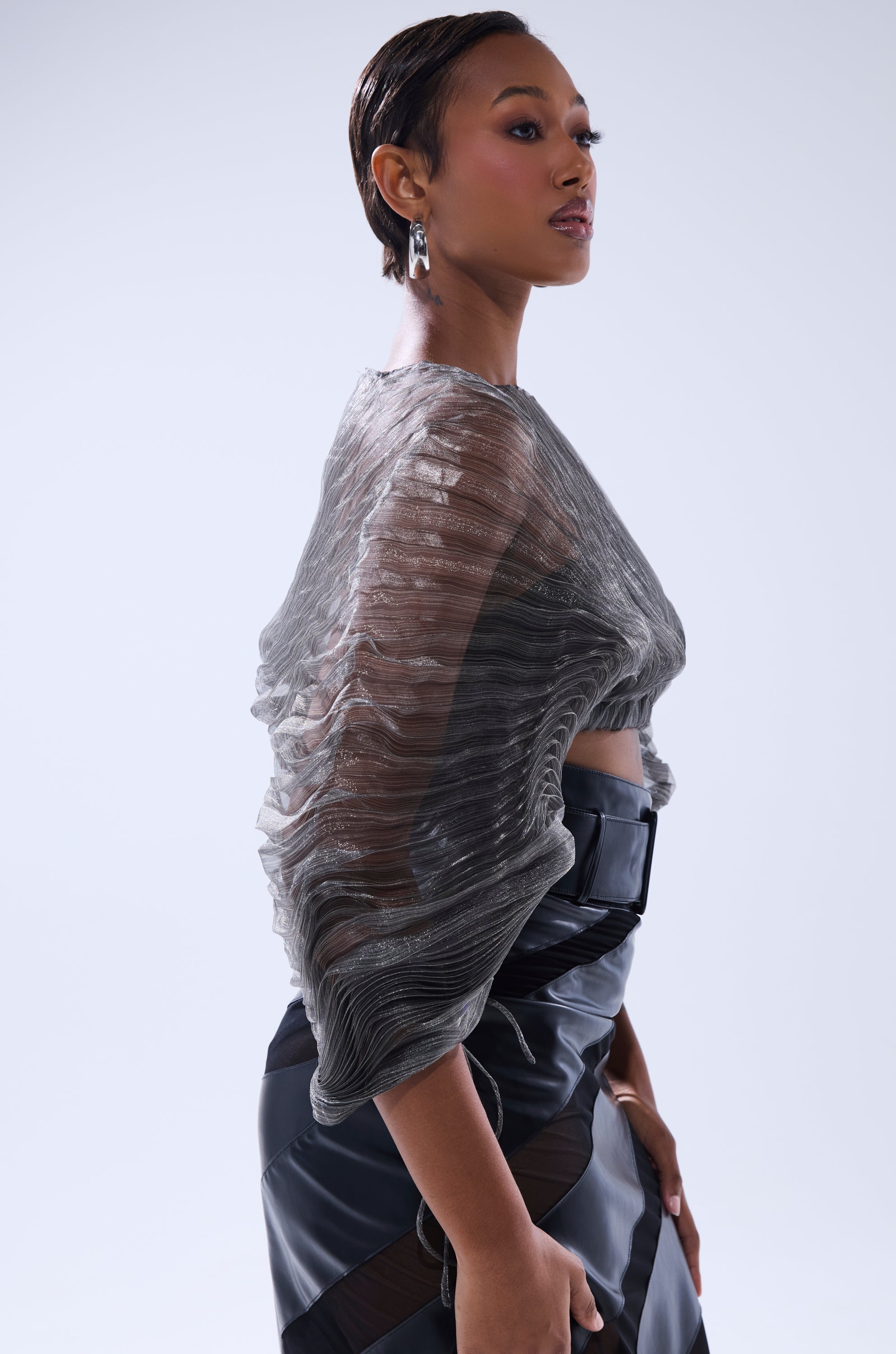 A woman models the SHEER PLEATED CROP TOP IN GREY with voluminous sleeves, styled with a black high-waisted belt, statement earrings, and short, sleek hair against a plain light background. 3