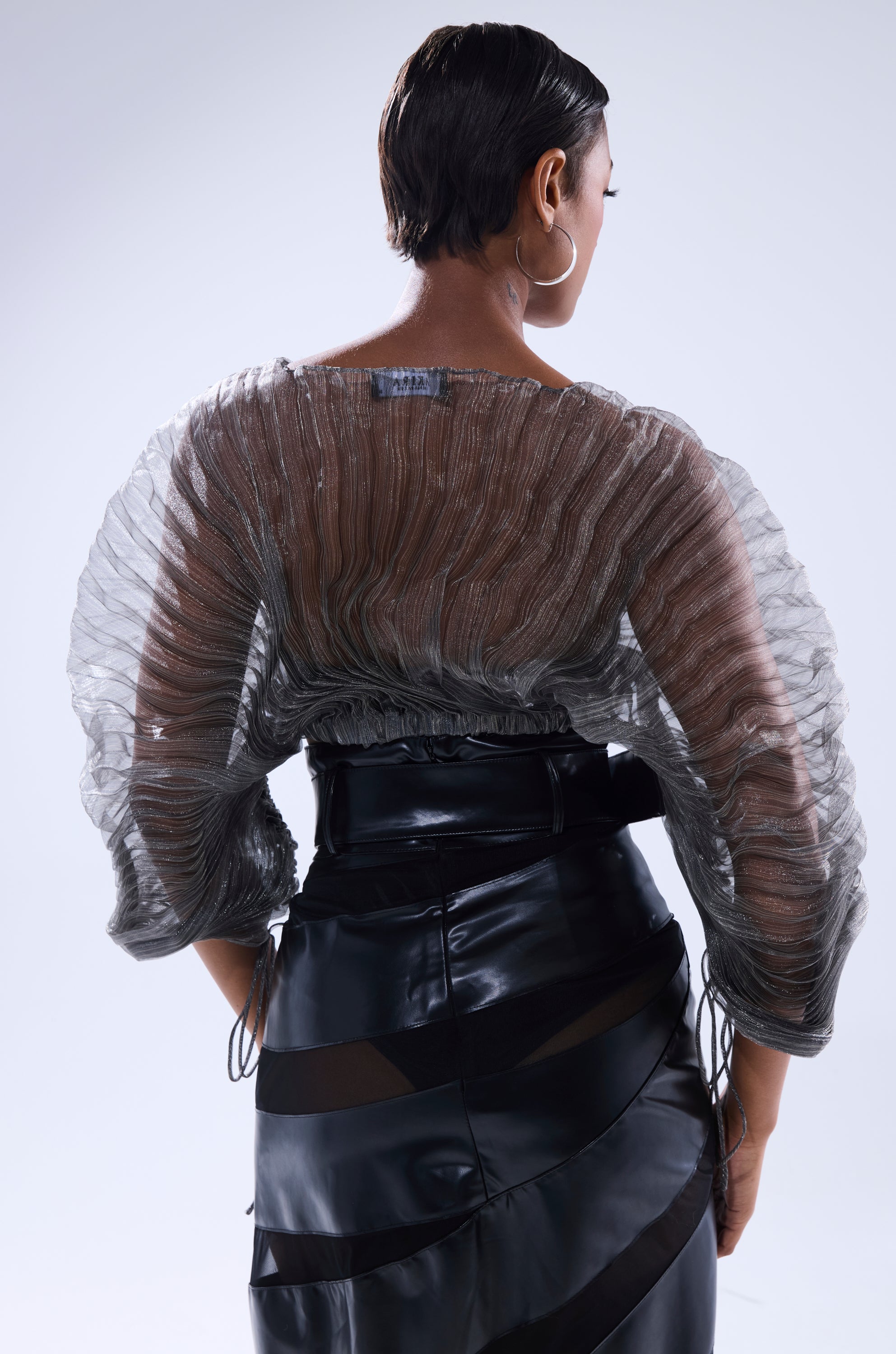 A person with short hair is seen from behind, wearing the SHEER PLEATED CROP TOP IN GREY and a high-waisted, glossy black skirt with sheer panels. 4