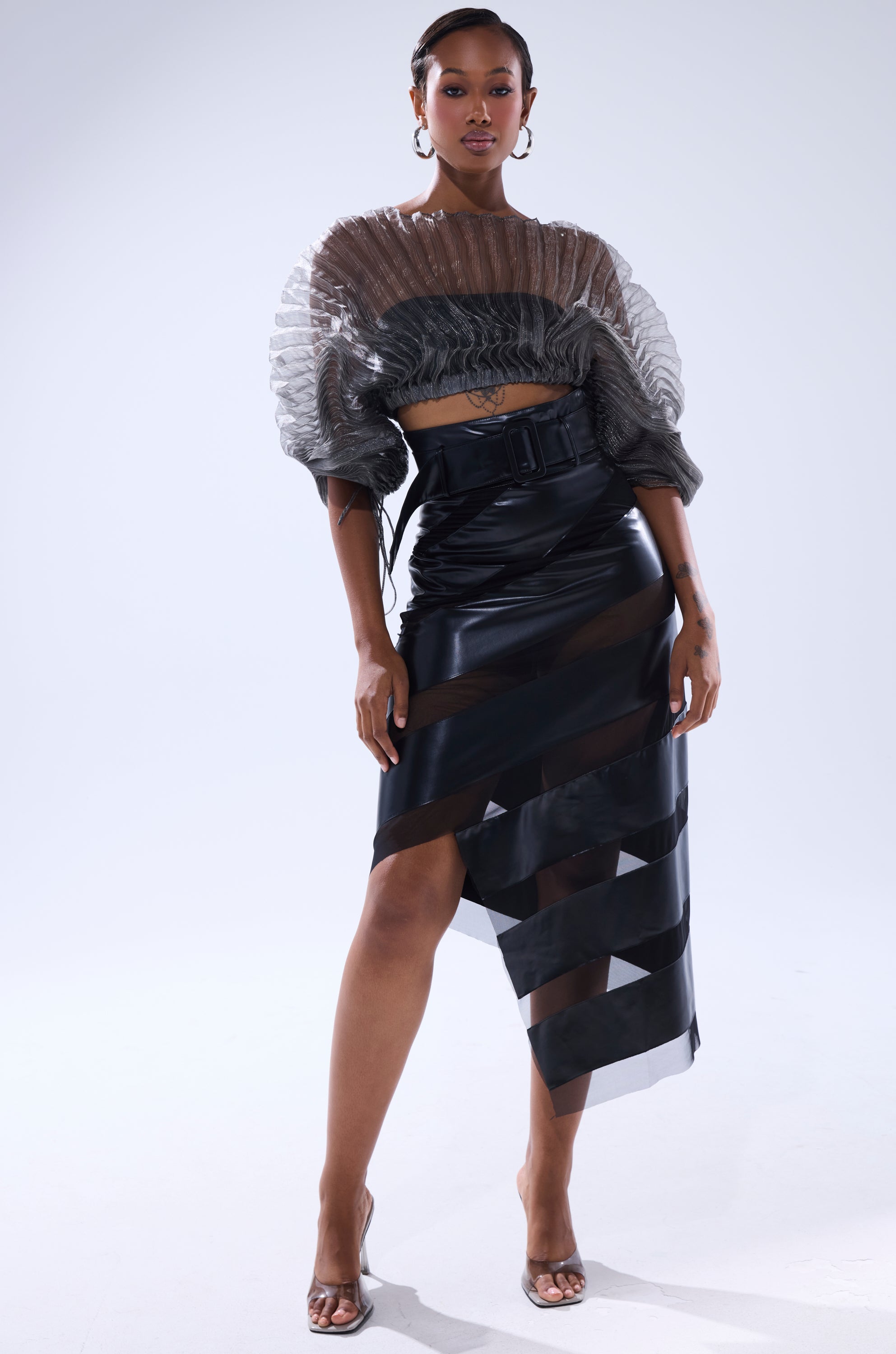 A woman poses in the SHEER PLEATED CROP TOP IN GREY with a high-waisted, asymmetrical black skirt featuring sheer panels. She accessorizes with clear heels and hoop earrings, her hair styled back, against a plain light background. 5
