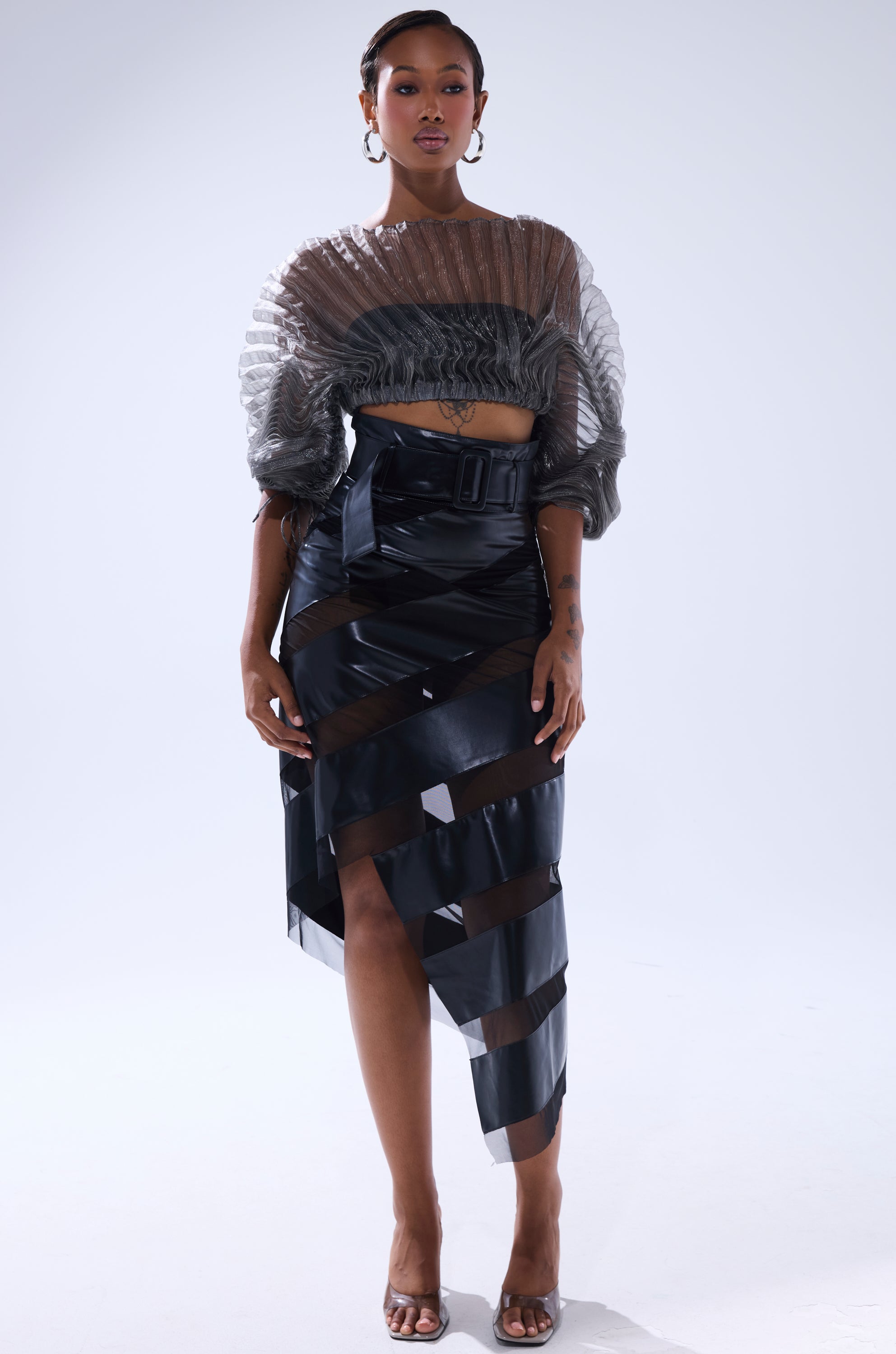 A woman stands confidently in the SHEER PLEATED CROP TOP IN GREY, featuring puffed sleeves. She pairs it with a high-waisted black skirt with wide sheer cutouts, accessorizing with hoop earrings and open-toe heels. 6