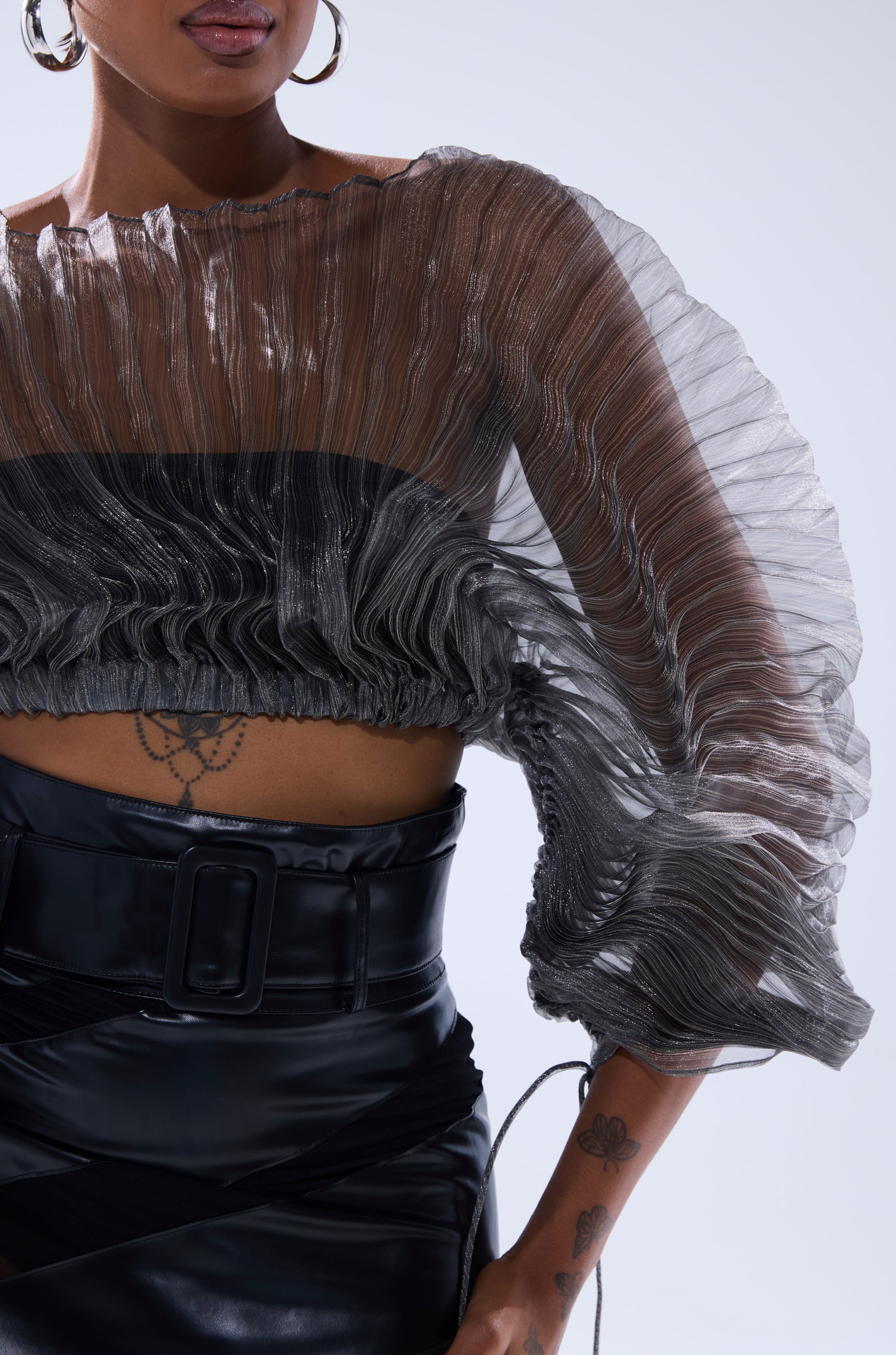 Woman wearing the SHEER PLEATED CROP TOP IN GREY with a high-waisted black skirt and belt; tattoos visible on torso and arm; accessorized with large hoop earrings. Photo shows her from shoulders to thighs. 7