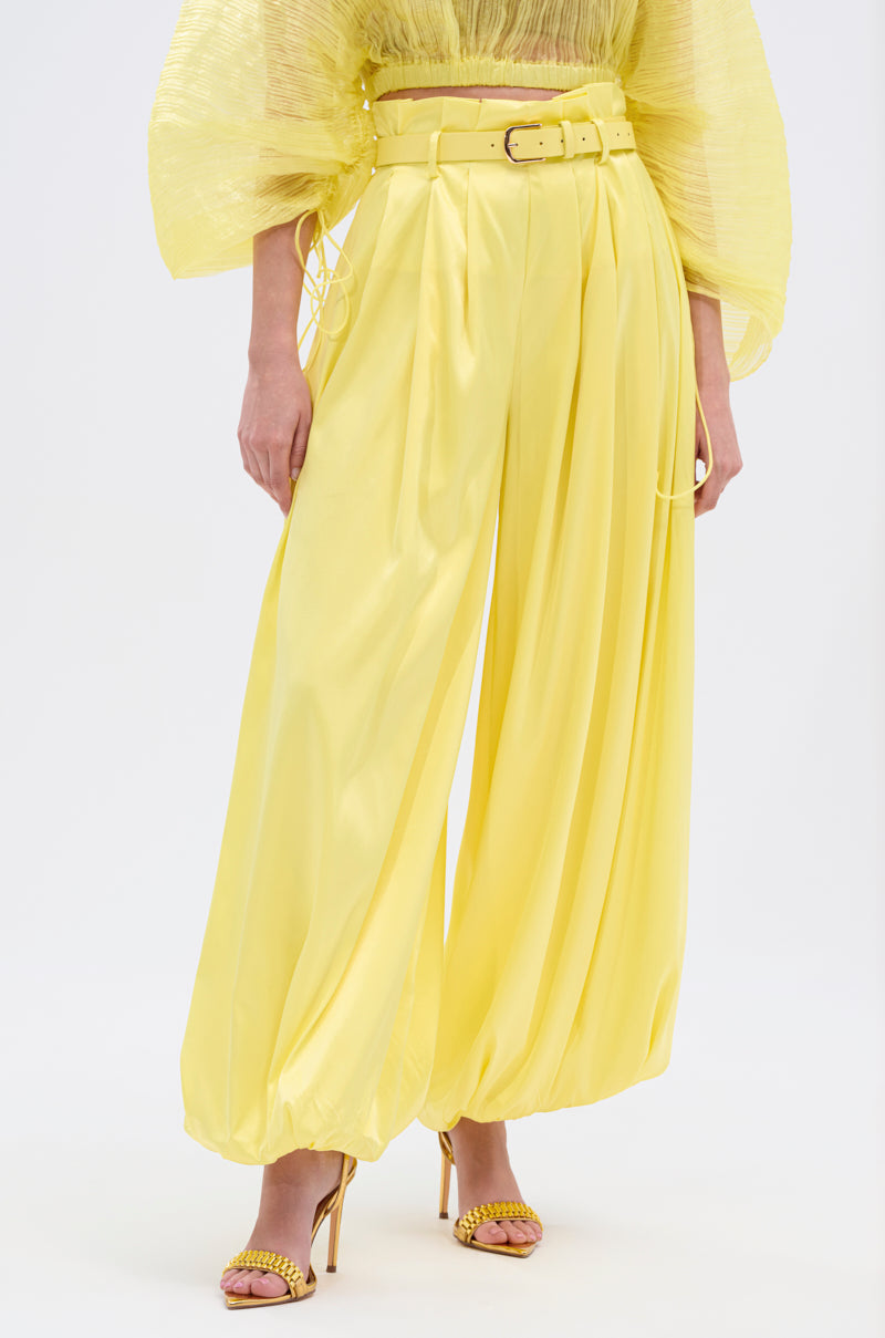 A person in the FEELING WILD BELTED BALLOON PANT—vivid yellow, high-rise, satin with a belt—paired with a sheer yellow voluminous-sleeved top and matching heels, stands against a plain white background.
