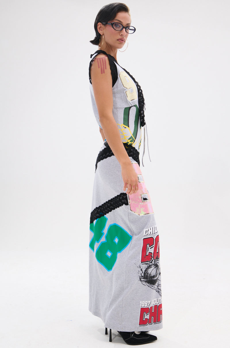 With short dark hair and glasses, a person stands sideways in the INSTA SAW IT FIRST GRAPHIC MAXI DRESS—featuring sporty-themed patchwork prints, bold numbers, and black braided details—set against a plain white background.