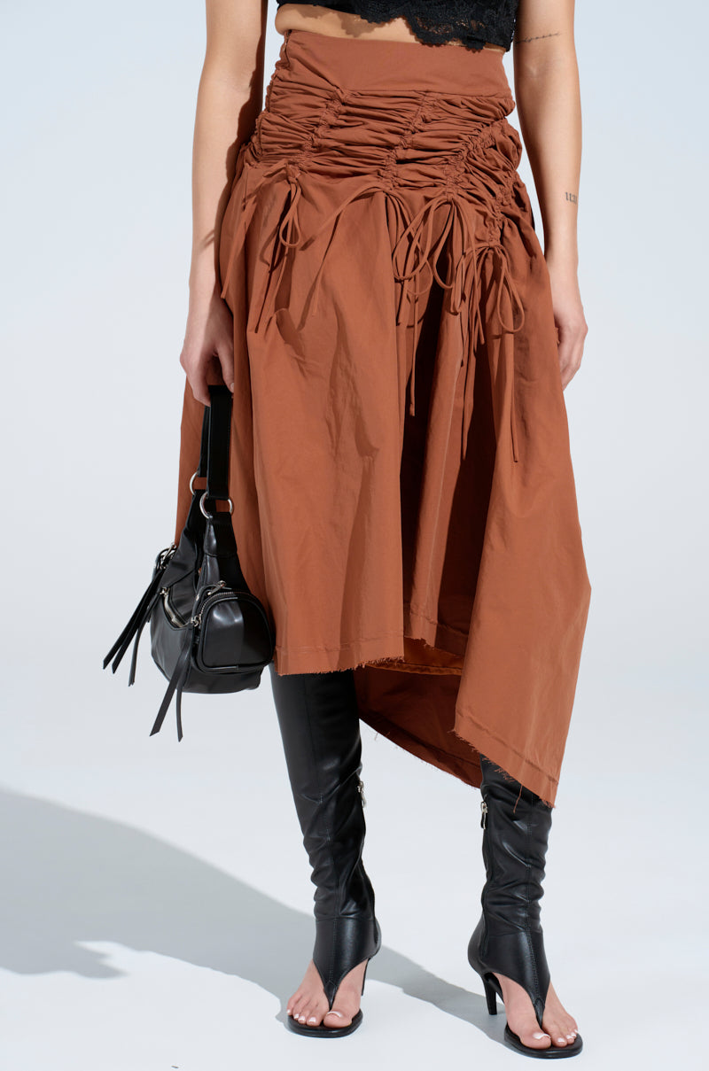 Wearing the GET WITH IT MAXI SKIRT, an asymmetrical brown skirt with ruched panels, paired with black over-the-knee open-toe boots and a sleek black handbag against a plain white backdrop to let the stylish ensemble shine.