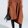 Wearing the GET WITH IT MAXI SKIRT, an asymmetrical brown skirt with ruched panels, paired with black over-the-knee open-toe boots and a sleek black handbag against a plain white backdrop to let the stylish ensemble shine.