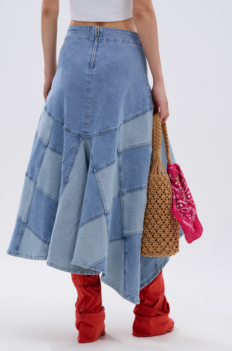 A person wears the ALL WE NEED IS LOVE DENIM MIDI SKIRT with a white crop top and red knee-high boots, holding a woven straw bag decorated with a pink bandana. The image is captured from behind against a plain background.