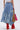 A person wears the ALL WE NEED IS LOVE DENIM MIDI SKIRT with a white crop top and red knee-high boots, holding a woven straw bag decorated with a pink bandana. The image is captured from behind against a plain background.