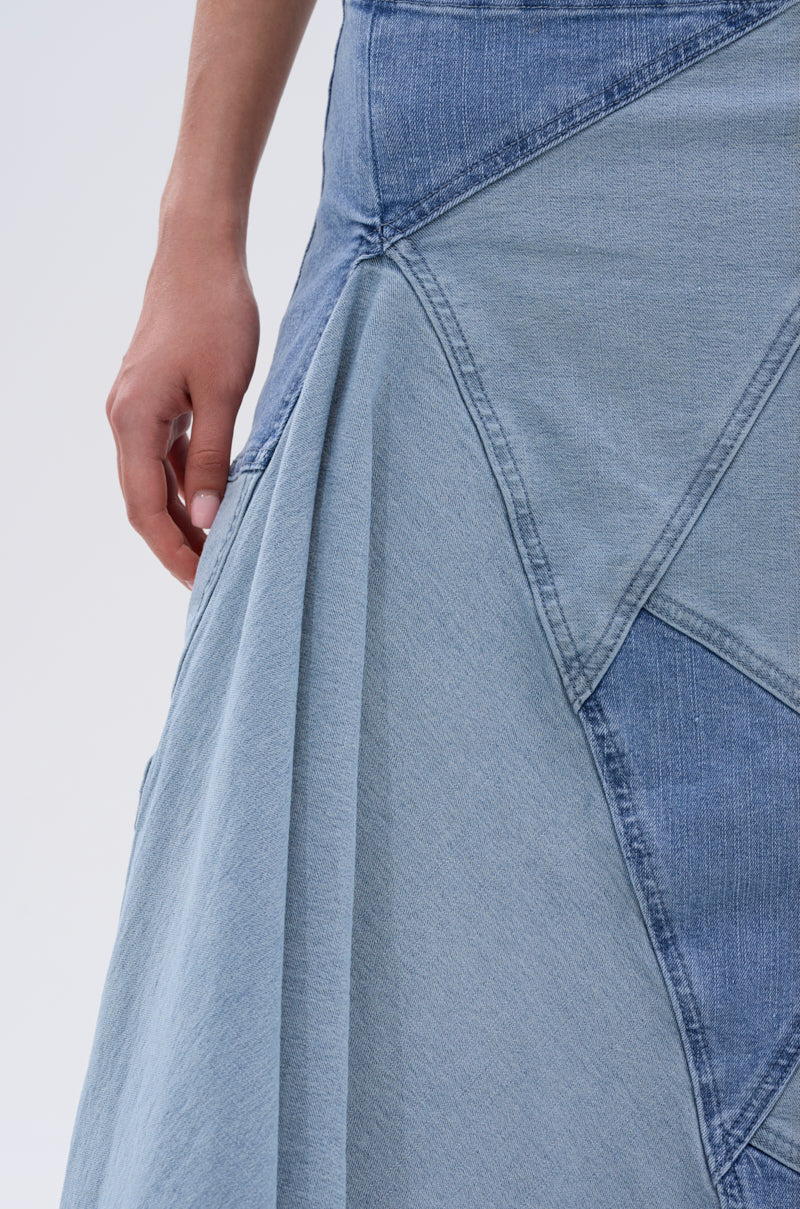 A close-up of a person wearing the ALL WE NEED IS LOVE DENIM MIDI SKIRT, featuring a high-rise fit and patchwork in various blue shades with triangular and paneled stitching, their left hand relaxed by their side.
