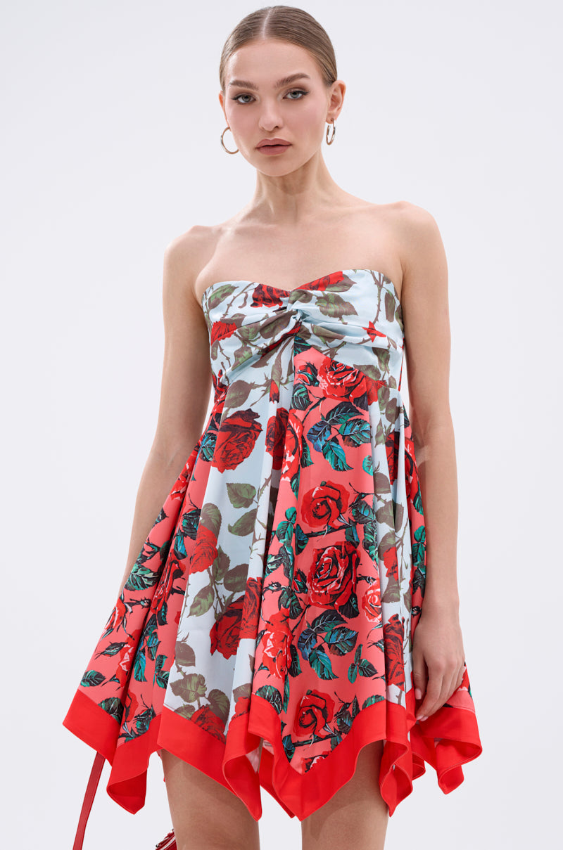 A woman stands against a plain light background wearing the ON VACAY PRINTED SATIN MINI DRESS, which features a strapless sweetheart neckline, handkerchief hem, bold red floral print with red trim. She accessorizes with hoop earrings and has her hair pulled back.