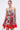 A woman stands against a plain light background wearing the ON VACAY PRINTED SATIN MINI DRESS, which features a strapless sweetheart neckline, handkerchief hem, bold red floral print with red trim. She accessorizes with hoop earrings and has her hair pulled back.