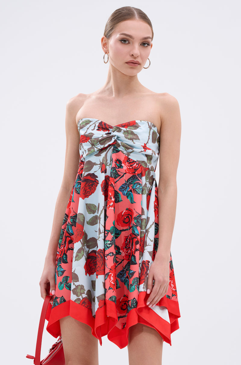 A woman in the ON VACAY PRINTED SATIN MINI DRESS, featuring a strapless sweetheart neckline, red and green rose print, and handkerchief hem, stands against a plain background holding a red handbag.