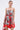 A woman in the ON VACAY PRINTED SATIN MINI DRESS, featuring a strapless sweetheart neckline, red and green rose print, and handkerchief hem, stands against a plain background holding a red handbag.