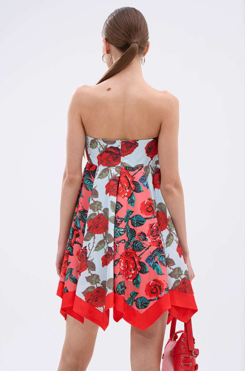 A woman with straight brown hair in a low ponytail models the ON VACAY PRINTED SATIN MINI DRESS, featuring a strapless sweetheart neckline, red and green rose print, handkerchief hem, and holds a red handbag. She is shown from the back against a light background.