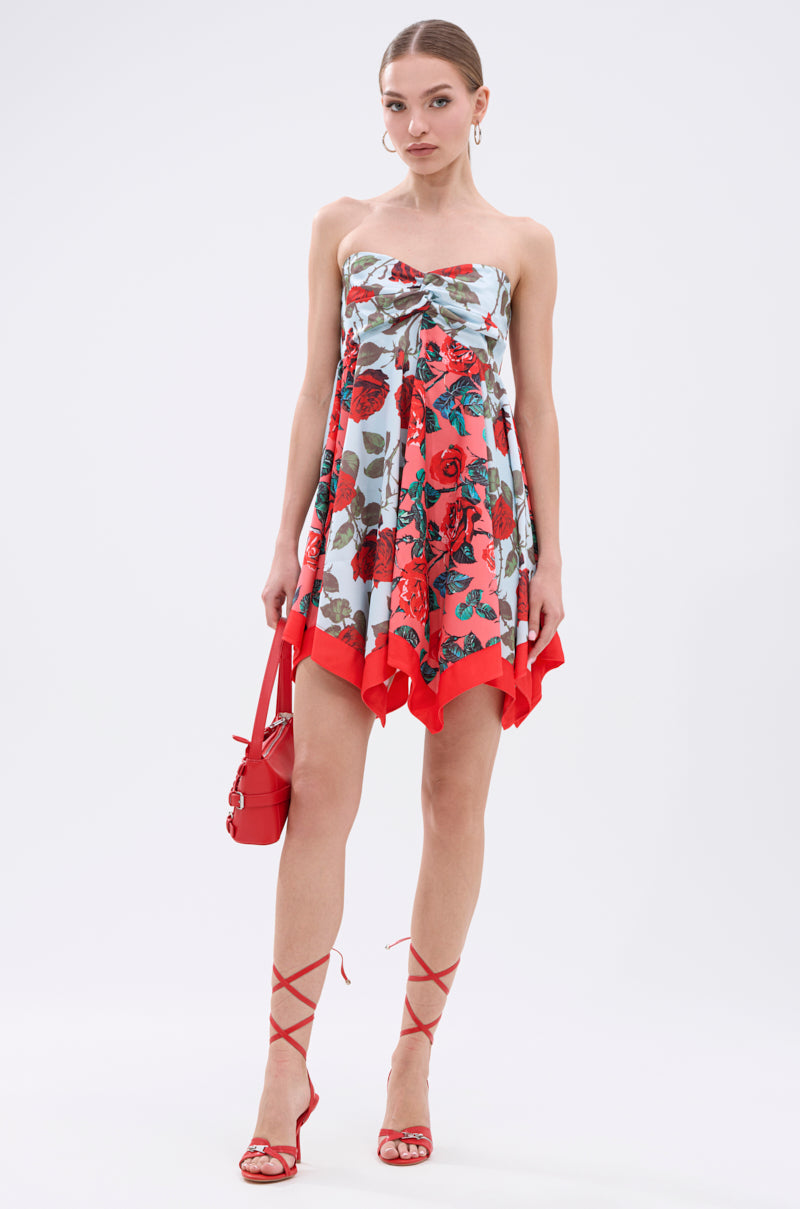 A woman stands against a plain white background wearing the ON VACAY PRINTED SATIN MINI DRESS with a strapless sweetheart neckline, handkerchief hem, red, white, and green rose print, paired with red lace-up heels and a red handbag.