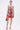 A woman stands against a plain white background wearing the ON VACAY PRINTED SATIN MINI DRESS with a strapless sweetheart neckline, handkerchief hem, red, white, and green rose print, paired with red lace-up heels and a red handbag.