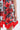 A close-up of a person wearing the ON VACAY PRINTED SATIN MINI DRESS, featuring a vibrant red rose print, green leaf accents, and a red-bordered asymmetric hem. The person's arm rests naturally at their side.