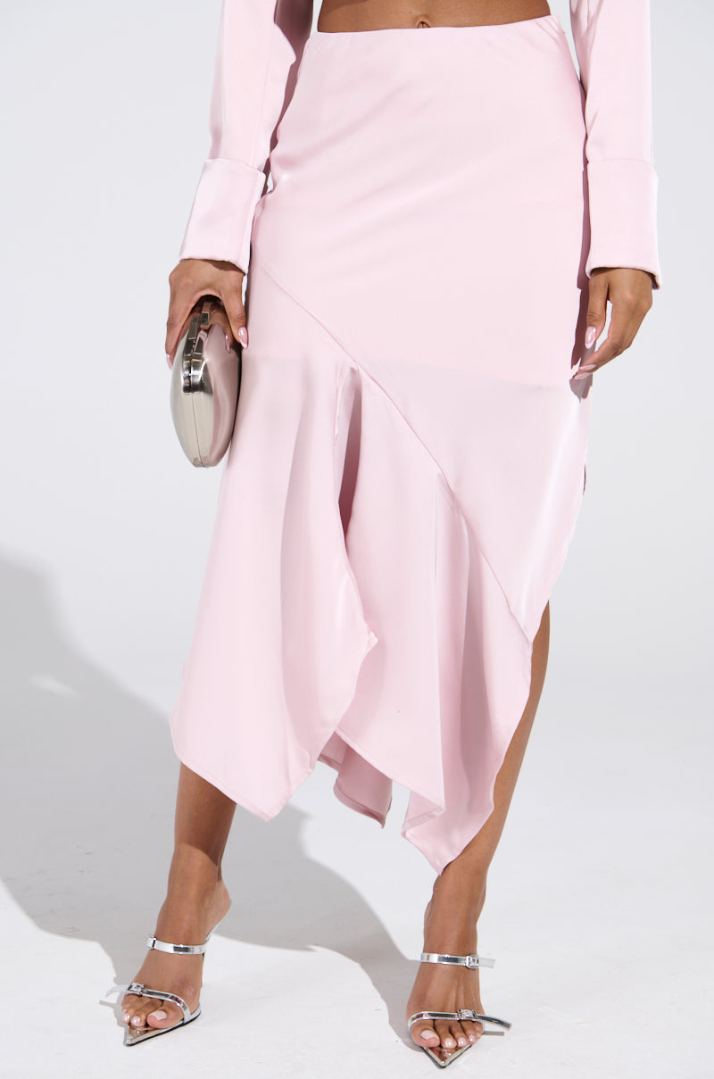 A person in a light pink SOFIA SATIN MIDI SKIRT, high-rise with an asymmetrical cut and high slit, pairs it with long sleeves. They clutch a metallic purse and step into silver strappy heels against a plain white backdrop.