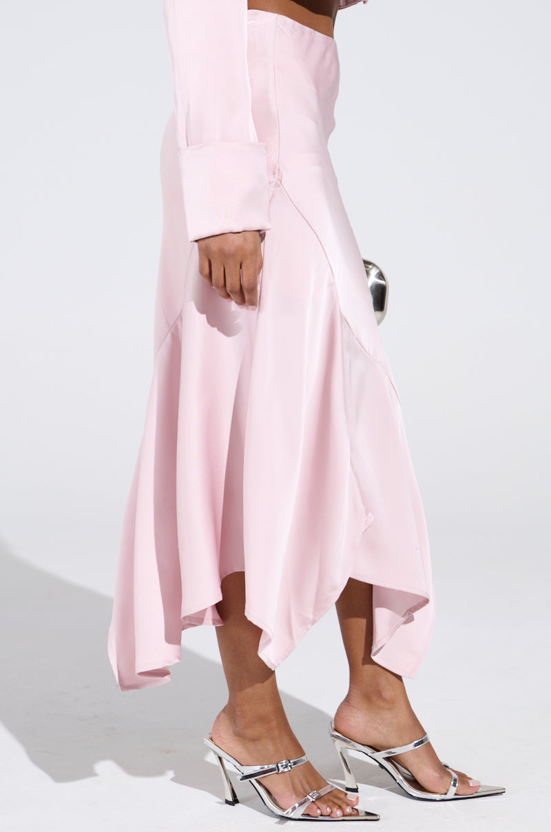 Person wearing the SOFIA SATIN MIDI SKIRT in light pink, paired with a long-sleeve top and silver high-heeled sandals. The high-rise skirt features a flowing, asymmetrical hemline against a plain white background.