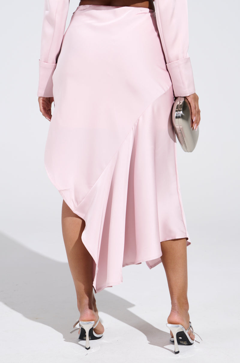 In a SOFIA SATIN MIDI SKIRT with an elegant asymmetrical hemline, a person in pastel pink stands on a white background. They hold a silver clutch, matching their long-sleeved top and highlighting open-toed high heels with ankle straps.