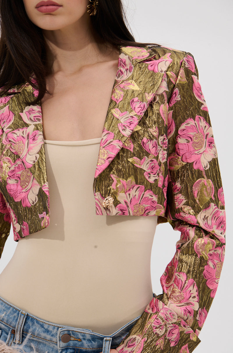 A woman wears the LIGHT THE WAY BROCADE BLAZER, featuring gold fabric with pink floral patterns and metallic details, styled over a beige bodysuit and light blue jeans. Her dark hair falls over her shoulders, with her face partly out of frame. 9
