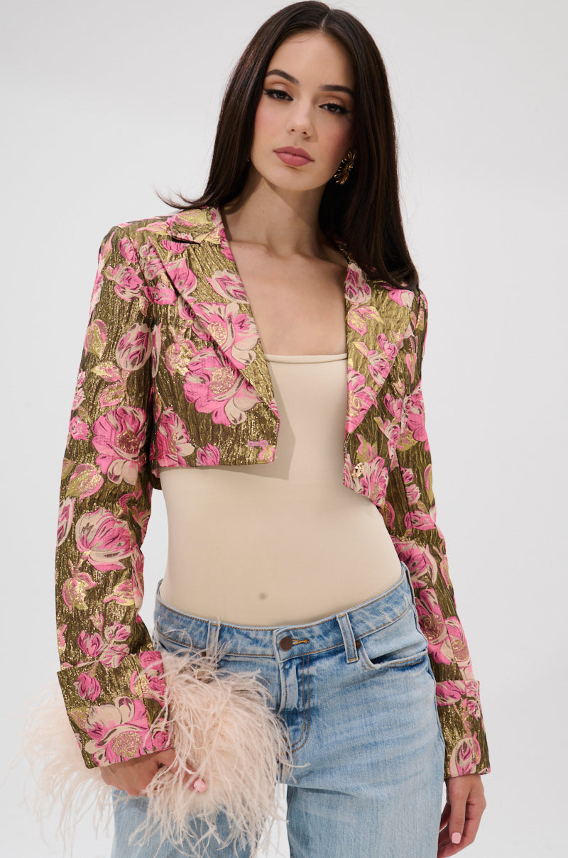 A woman with long brown hair models the LIGHT THE WAY BROCADE BLAZER—gold and pink with metallic details—over a beige top and light blue jeans, holding a bag with pink feather accents, against a plain white background. 1