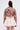 A person stands facing away, wearing the LIGHT THE WAY BROCADE BLAZER—a pink floral jacket with metallic details—and a light beige flared skirt. Their hair is partially tied up with large hoop earrings, set against a plain white background.