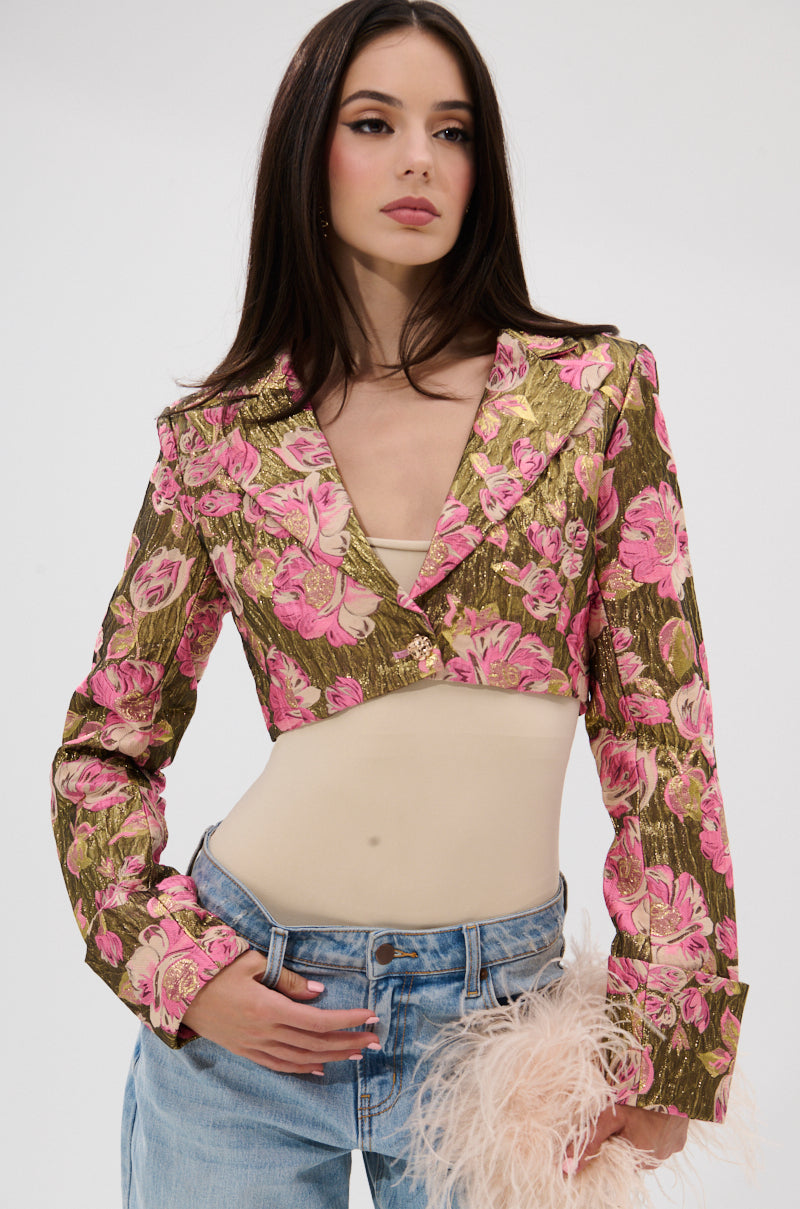 Model poses against a plain light background wearing the LIGHT THE WAY BROCADE BLAZER in pink and gold with metallic details, styled over a beige top, blue jeans, and holding a light pink feathered accessory. 2