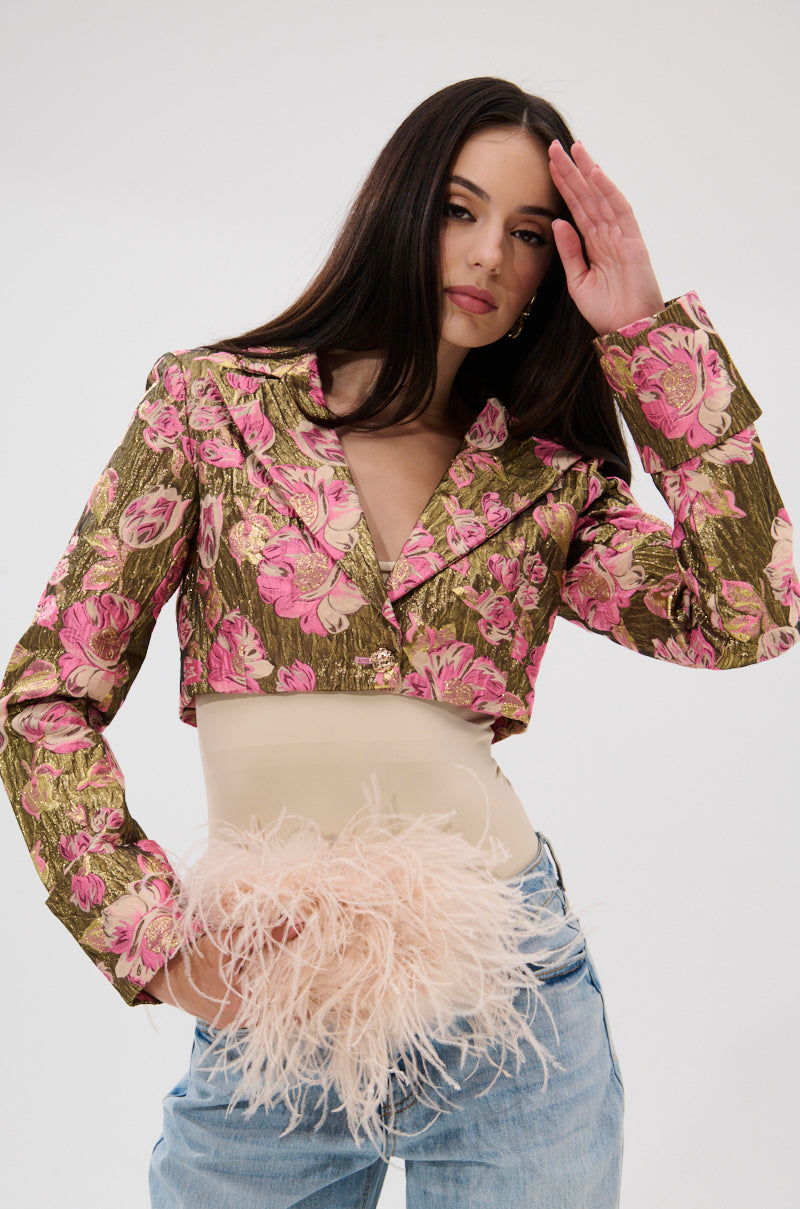 A woman with long dark hair wears the LIGHT THE WAY BROCADE BLAZER, a cropped fitted piece with pink and gold floral patterns, paired with light blue jeans. She poses against a plain white background holding pale pink feathers at her waist. 3