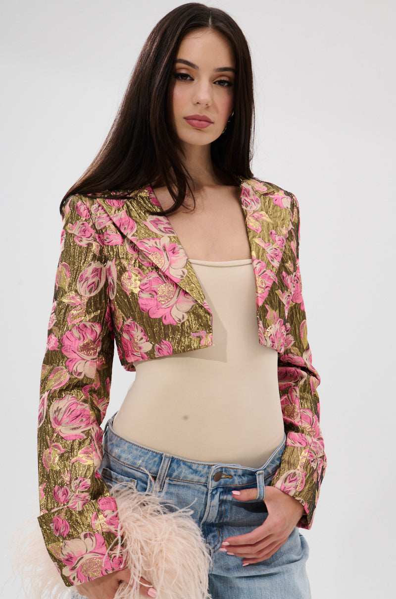 A woman with long dark hair models the LIGHT THE WAY BROCADE BLAZER—gold and pink with metallic details—over a beige top and blue jeans, holding a pale pink feathered accessory against a plain light background. 4