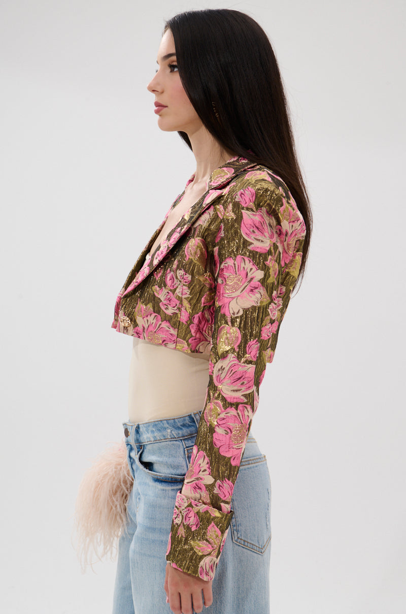 Woman in profile against a plain background, wearing the LIGHT THE WAY BROCADE BLAZER with pink floral patterns over a beige top, light blue jeans, and a fluffy beige accessory at her waist. 5
