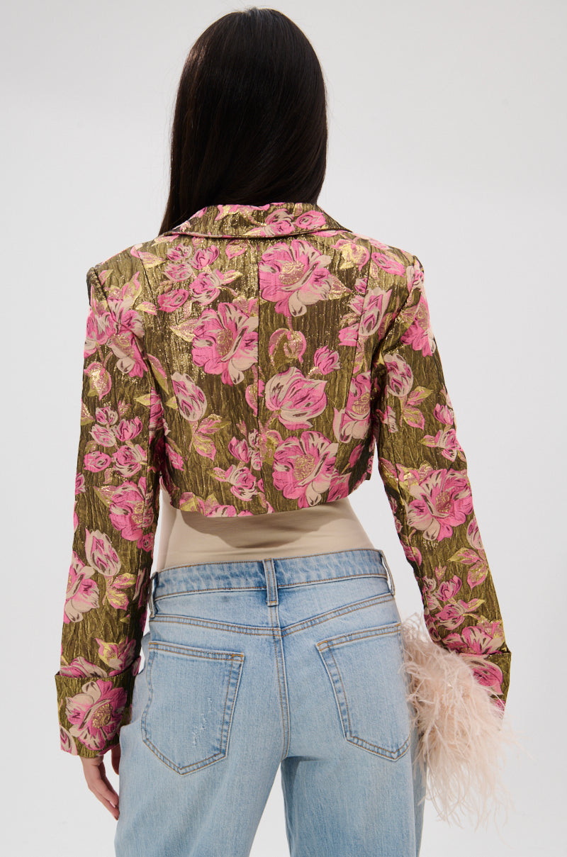 A person with long dark hair, shown from the back against a plain light background, wears the LIGHT THE WAY BROCADE BLAZER—gold and pink with a cropped silhouette—over a beige top and light blue jeans, holding a beige feathered accessory. 6