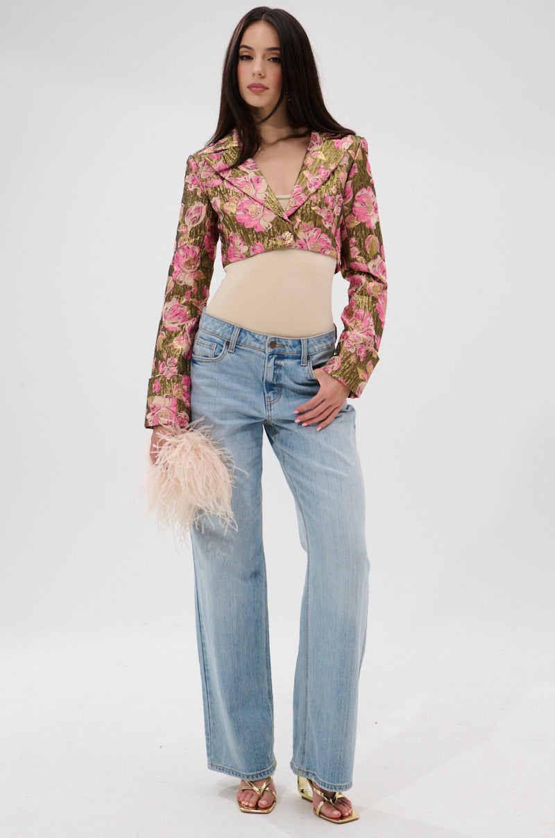 A woman stands against a plain background wearing the LIGHT THE WAY BROCADE BLAZER, a beige top, light blue wide-leg jeans, gold sandals, and holding a pale pink feathered clutch. 8