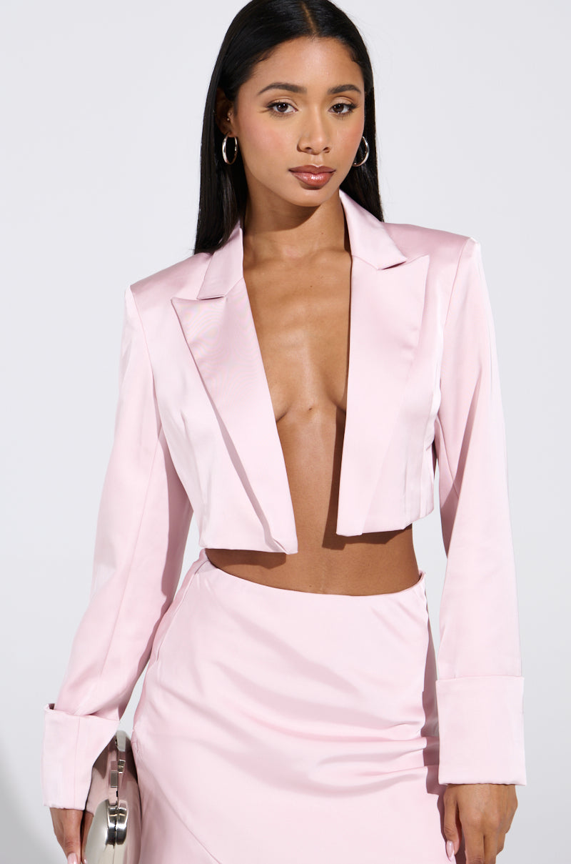 A woman poses confidently in the SOFIA CROPPED SATIN BLAZER—light pink with a notch lapel collar—and a matching skirt, holding a silver clutch. She wears hoop earrings as her long dark hair flows against the plain white background.