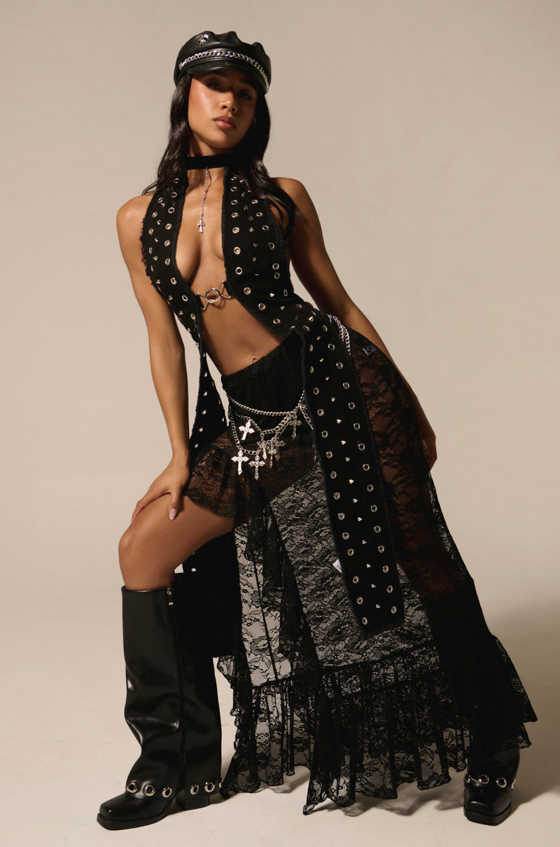 A woman poses in a black, gothic outfit with a black 'Dance the Night Away Lace Maxi Skirt,' boots, and a hat. The dress, detailed with metal eyelets and accessorized with chains and crosses, offers a festival-ready look against the neutral studio backdrop.