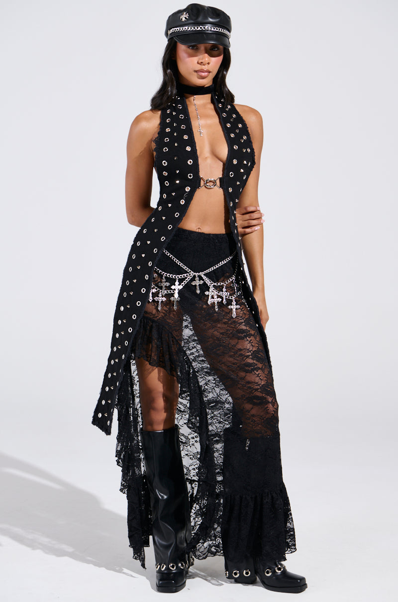 A person radiates a festival-ready vibe in a sleeveless black outfit with grommet accents and the DANCE THE NIGHT AWAY LACE MAXI SKIRT. Tall boots complement the ensemble, accessorized by a black cap, silver chain belt with cross charms, and choker. Their confident stance showcases their style.