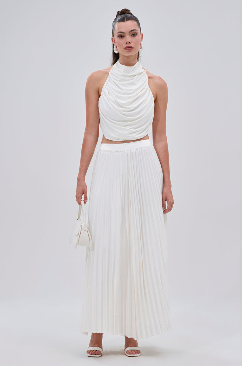 A woman poses against a plain background in the SWEET THING PLEATED SATIN MAXI SKIRT, styled with a sleeveless white halter top, white sandals, a small white handbag, and her hair up in a high ponytail. 1